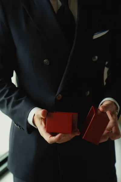Wedding ring box held by groom holding ring box, a red case against a navy double-breasted suit in soft window light indoors