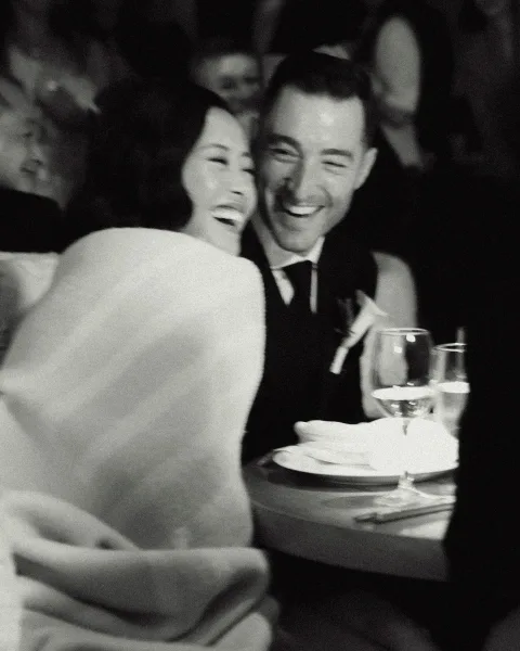 Reception couple portrait in a black and white wedding photo, bride and groom laughing at their table with wine glasses in a dim banquet room