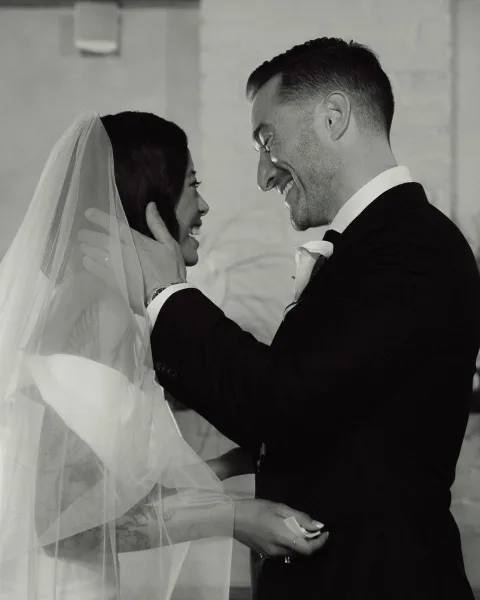 Wedding couple portrait in black and white showing groom holding bride’s face as they smile close together by a stone wall indoors