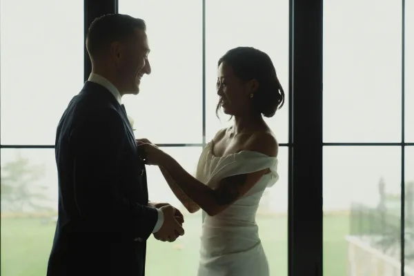 First look moment as the bride adjusts groom tie, off-the-shoulder dress and boutonniere in soft window light with landscape view