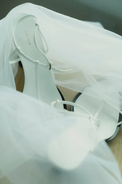 Bridal shoes in white wedding heels, strappy with ankle buckle, resting on a wood floor beside soft tulle veil folds