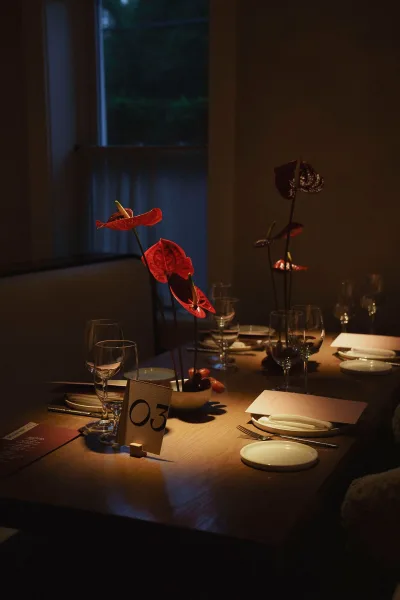 Reception tablescape with wedding table setting, red anthurium in bud vases, menus, place cards, and glassware on a wood table by a window