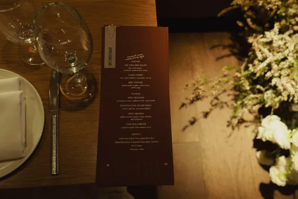 Wedding menu card clipped with a gold paperclip beside wine glasses, plate, knife, and folded napkin on a warm-lit wooden table with florals