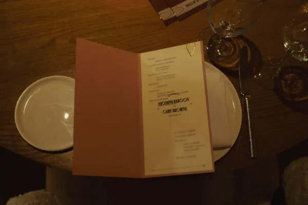 Wedding program with ceremony program design on a pink cover, clipped beside a plate, linen napkin, knife, and wine glass on a warm wood table