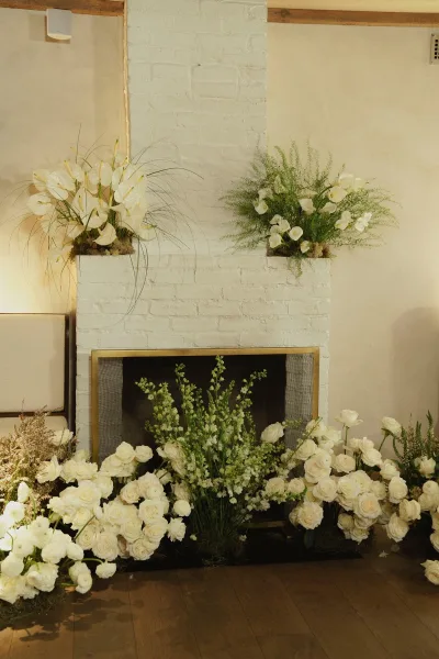 Ceremony backdrop decor with fireplace wedding altar, white roses, calla lilies, anthurium and greenery on a white brick wall indoors