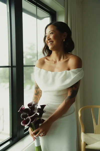 Bridal portrait of a bride holding bouquet, gazing out a large window in soft light, wearing an off-the-shoulder dress with tattoos visible