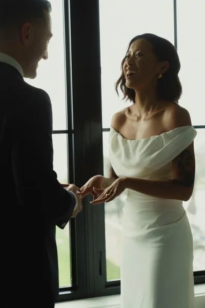 First look moment as bride and groom laugh by large windows, bride in off the shoulder wedding dress showing her ring in soft indoor light