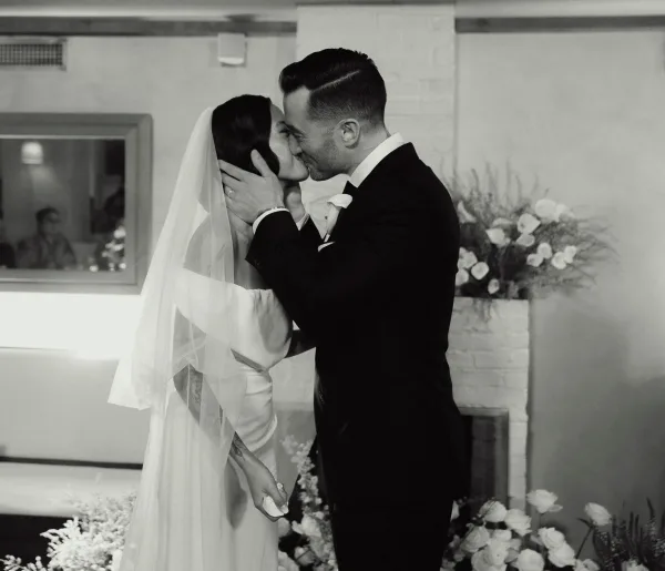 Wedding kiss in a black and white wedding photo, groom gently holding bride’s face as they kiss by a fireplace mantel with florals