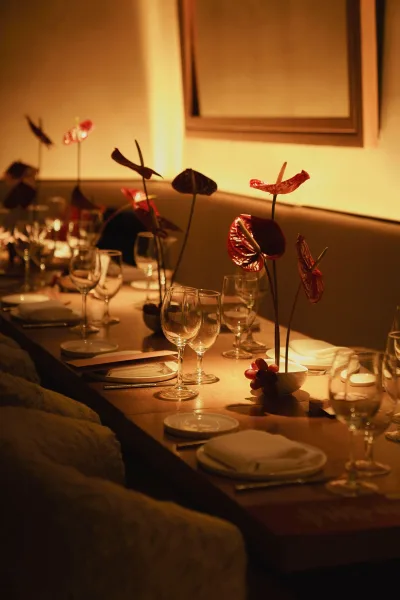Reception tablescape with long banquet table setting, red anthurium in bud vases, grapes, plates and glassware in a warmly lit restaurant interior