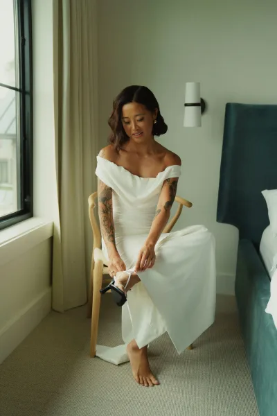 Bridal portrait of a bride putting on shoes in an off the shoulder wedding dress, seated on a wooden chair by hotel window light