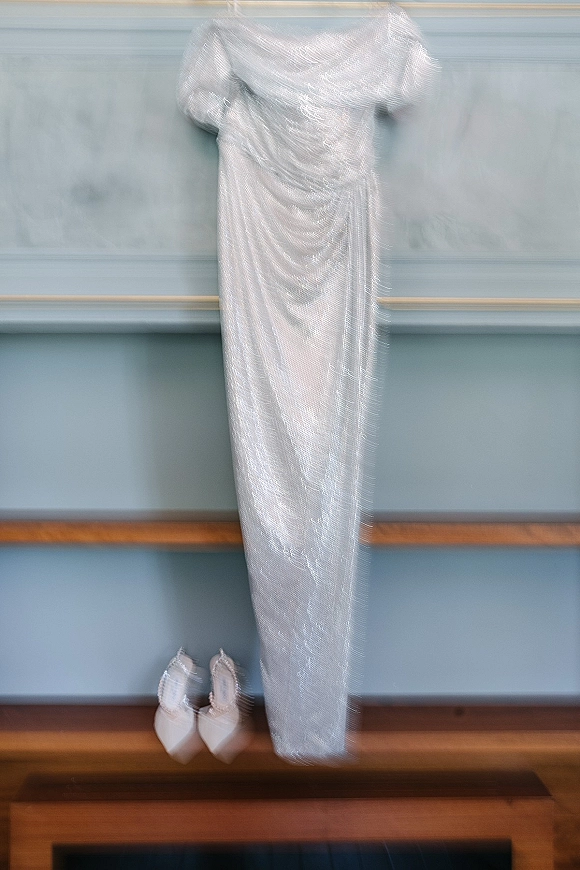 Wedding dress displayed with pointed toe bridal heels, featuring an off the shoulder sequin gown against a blue paneled wall and wooden bench
