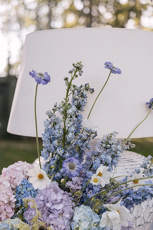 Wedding cake with fresh flower topper of blue delphinium, hydrangeas, and daisies on buttercream, set against outdoor greenery bokeh lights