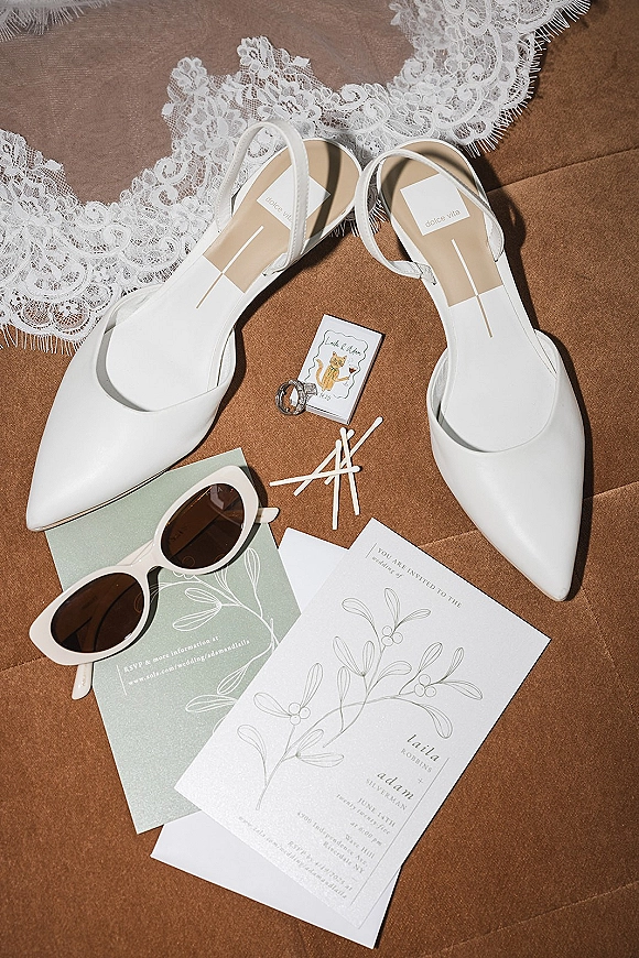 Wedding flat lay with wedding invitation suite, white bridal shoes, lace, rings, sunglasses, and matchbook on a brown fabric backdrop