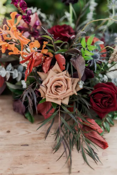 Bridal bouquet with burgundy wedding bouquet tones, roses and orchids with fern greenery, resting on a wood table with blurred greenery behind