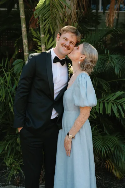 Couple portrait of bride kissing groom’s cheek, his black tuxedo bow tie and boutonniere against a palm leaf tropical backdrop