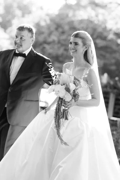 Wedding processional as bride walking down aisle holding a bridal bouquet, veil flowing beside a tuxedoed escort in a sunlit garden