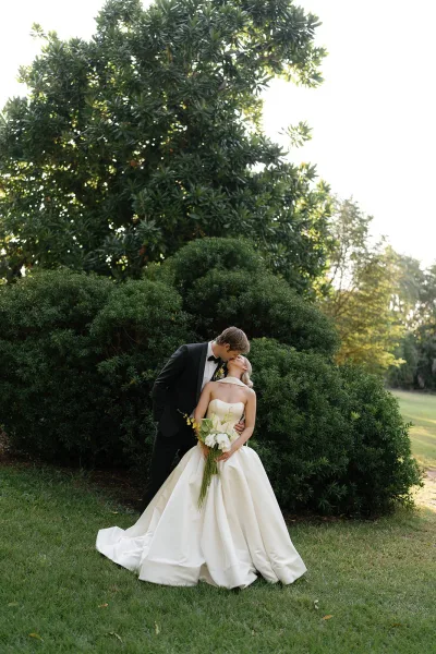 Wedding kiss portrait of bride and groom kissing, her strapless gown and calla lily bouquet beside his tuxedo, on a garden lawn