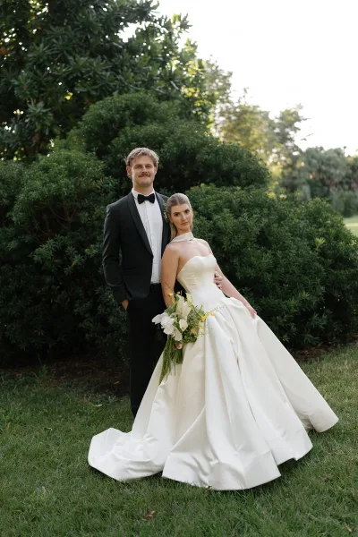 Couple portrait of bride in a strapless satin gown holding calla lilies and orchids beside groom in black tuxedo on a sunlit lawn