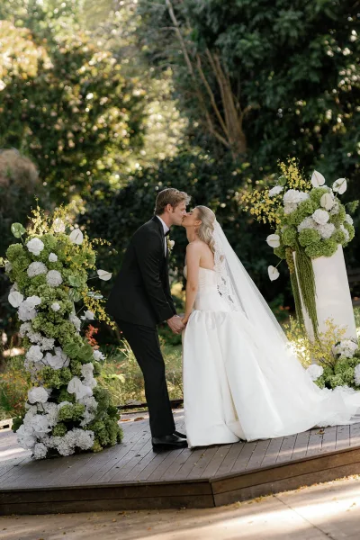 Wedding kiss as bride in strapless gown and long veil kisses groom in tuxedo beside floral pillars with white blooms and orchids on a garden platform