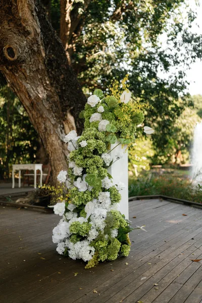 Wedding ceremony altar with floral ceremony pedestal of hydrangeas, white blooms, and yellow accents on a wooden deck beneath a large tree