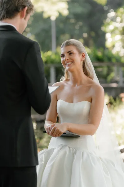 Ceremony moment as groom reads wedding vows from a vow book to a smiling bride in a strapless dress and veil in sunlit garden