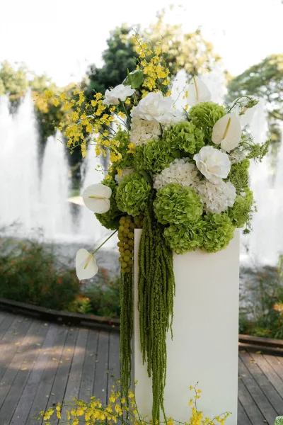 Wedding floral arrangement on a white pedestal plinth with green and white hydrangea, anthurium, yellow orchids, and cascading amaranthus by a garden fountain