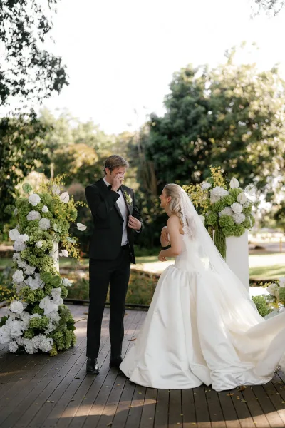 First look moment as bride in a ball gown and long veil approaches groom in black tuxedo on a sunny garden deck with white floral pedestals