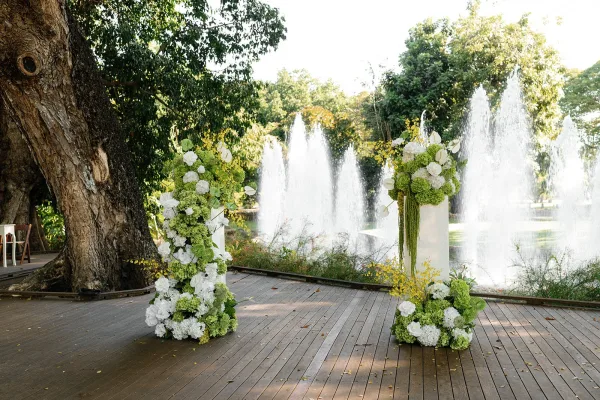 Ceremony altar decor with wedding altar flowers on pedestal columns and greenery, set on a wood deck by a lake with fountains and trees