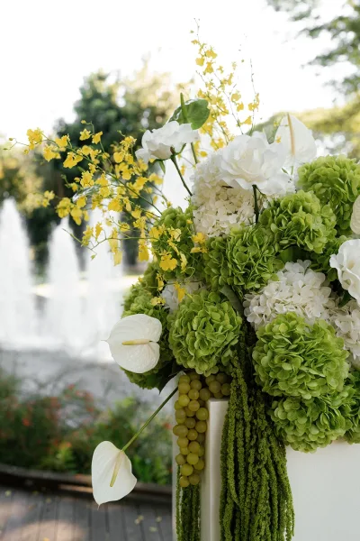 Wedding floral arrangement of green and white wedding flowers with hydrangeas and yellow orchid spray on a pedestal by a garden walkway and water feature