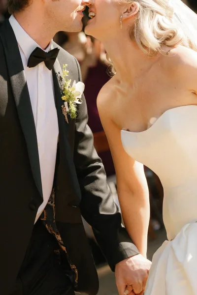 Wedding kiss as bride in strapless dress and veil kisses groom in tuxedo with black bow tie, guests blurred in sunlit outdoor ceremony