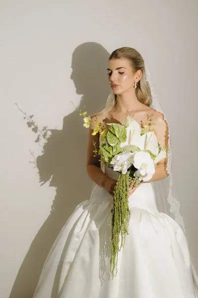 Bridal portrait of a bride in a strapless ball gown with cathedral veil, holding a white orchid bouquet with trailing greenery against a plain wall shadow