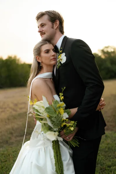 Couple portrait of bride and groom embrace as she holds a calla lily bouquet, posing in a field with trees in warm sunset light