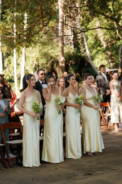 Bridesmaid lineup in champagne satin dresses holding white and greenery bouquets at a sunlit garden ceremony with guests and wooden chairs