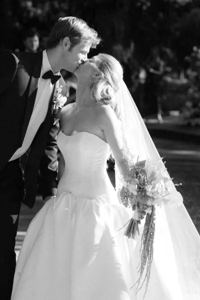 Wedding kiss in a black and white wedding photo as bride in strapless gown and veil kisses groom in tux, guests blurred in sunlit garden