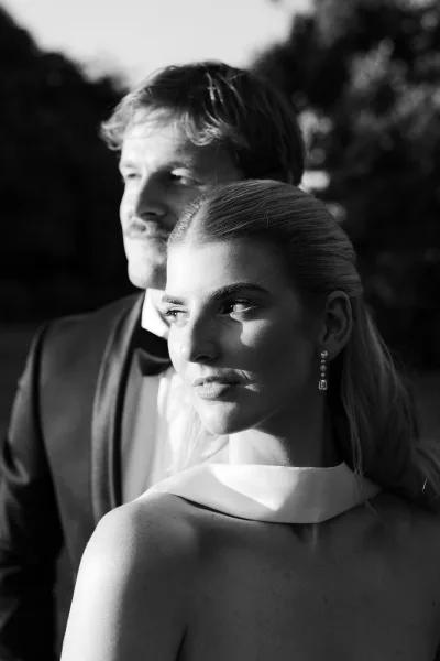 Couple portrait in black and white wedding portrait style, groom in tuxedo behind bride in high-neck dress, trees and sky beyond