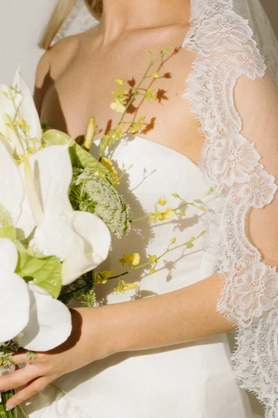 Bridal bouquet close-up with a white calla lily bouquet, yellow blooms and greenery held by a bride in lace sleeves against soft sunlight shadows
