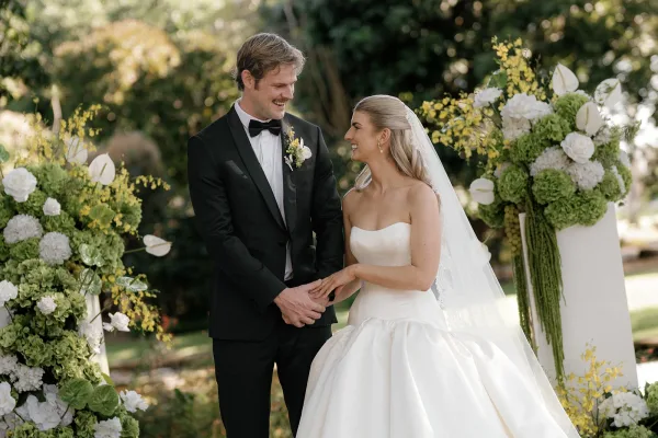 Couple portrait of bride and groom holding hands, veil flowing as she looks at him beside hydrangea florals in a garden setting