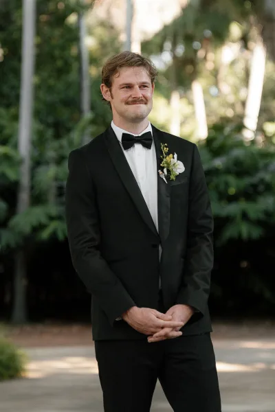 Groom portrait of a groom in tuxedo with black tuxedo, bow tie, and boutonniere, standing on a walkway with trees and greenery behind him
