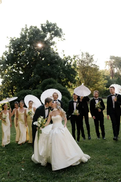 Wedding party photo of bride and groom kissing, walking with bridesmaids in gold and groomsmen in tuxedos on a sunny lawn with white parasols