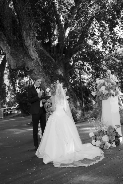Wedding vows as groom reading from a vow book into a microphone beside bride in lace veil on a wooden deck under a large tree