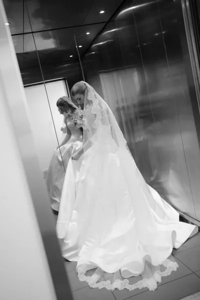 Bridal portrait in an elevator, bride looking down in a long wedding dress with lace veil and bouquet, reflected in mirrored wall