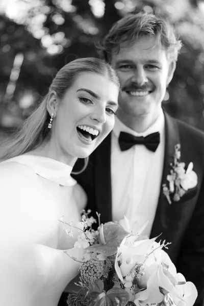 Couple portrait in a black and white wedding portrait, bride laughing over her shoulder beside groom in tuxedo with calla lily bouquet and trees behind