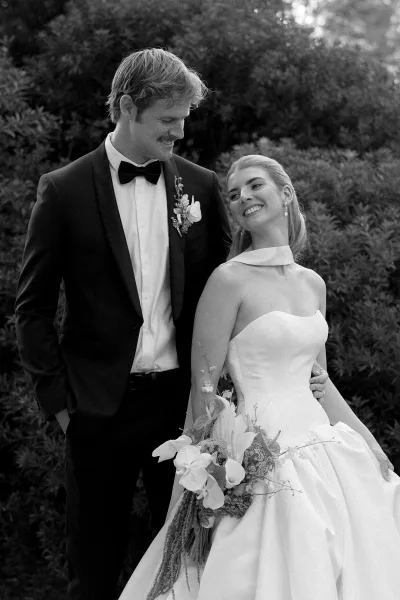 Couple portrait in a black and white wedding portrait style, bride smiling in strapless gown holding cascading bouquet as groom looks at her in garden greenery