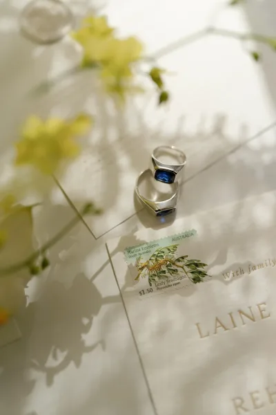 Wedding rings flatlay with silver bands and a blue gemstone ring on vellum invitation, envelope and vintage stamp, with yellow flowers on white paper