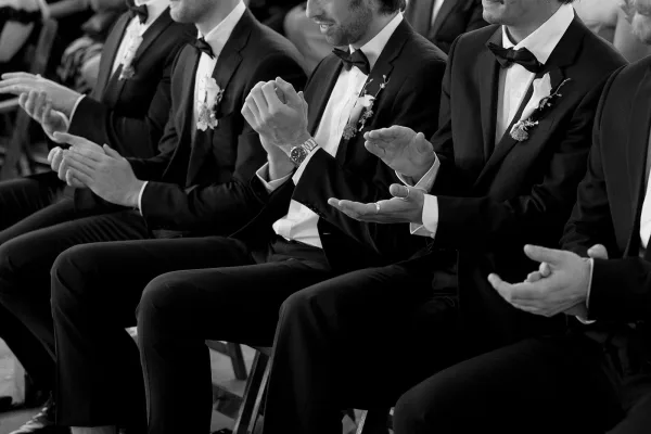 Groomsmen ceremony moment as men in black tuxedos applaud with boutonnieres and bow ties, with seated guests and chairs behind them