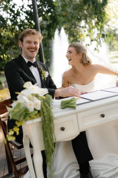 Wedding signing as bride in strapless dress and veil laughs beside groom in black tuxedo at patio table with white flowers and fountain