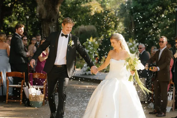 Wedding recessional as bride and groom walk the aisle holding hands, bouquet and veil flowing, guests tossing petals along a garden walkway