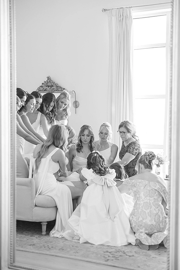 Bridesmaids getting ready in matching dresses, chatting by a bright window in a white bedroom, reflected in an ornate mirror frame