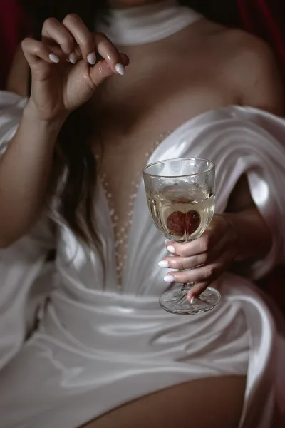 Bride with champagne in a satin bridal gown toasts with berries floating in the glass, set against a dark satin drape that frames the scene.
