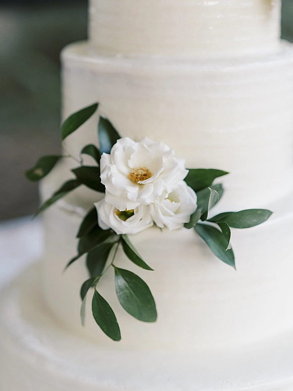 Wedding cake with smooth buttercream tiers topped with white flowers and greenery, styled against a neutral backdrop